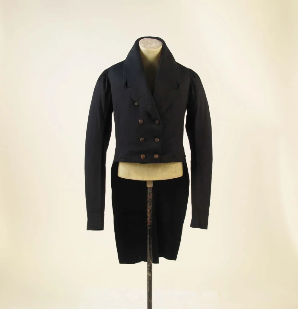 Historyczny męski frak z XVIII-XIX wieku: czarny, dwurzędowy surdut z wysokim kołnierzem, długimi klapami, guzikami, rozcięty z przodu, długi z tyłu z połami, wystawiony na manekinie.