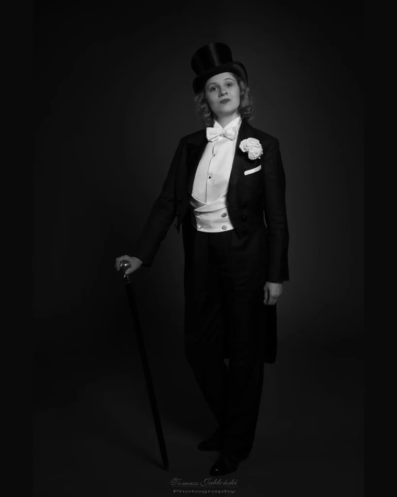 czarno biała fotografia Kobiety we fraku w stylu Marlnen Ditrich z laską w cylindrze i białymi różami w kieszonce