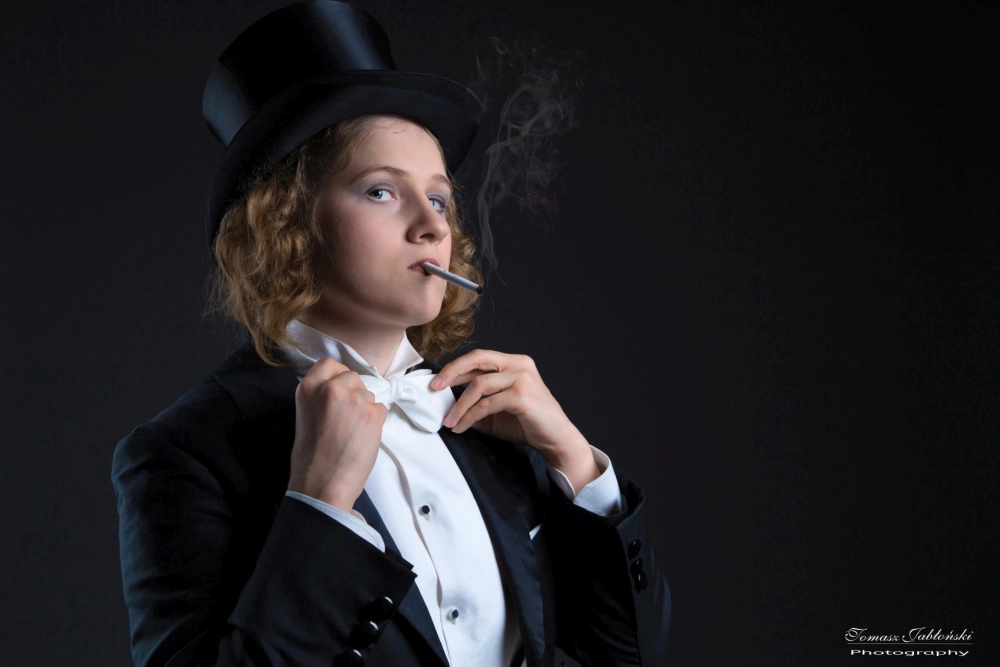 fotografia Kobiety we fraku i cylindrze w stylu Marlnen Ditrich z laską poprawiającą muszkę, pali papierosa.