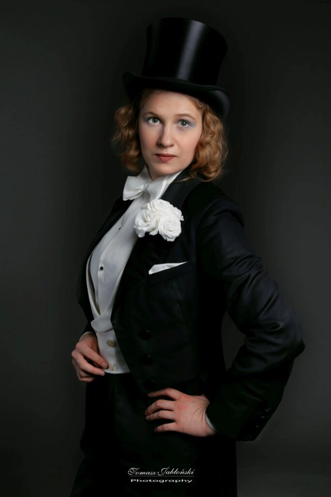 Fotografia Kobiety we fraku w stylu Marlnen Ditrich z dłonią na biodrze, w cylindrze z białymi różami w kieszonce